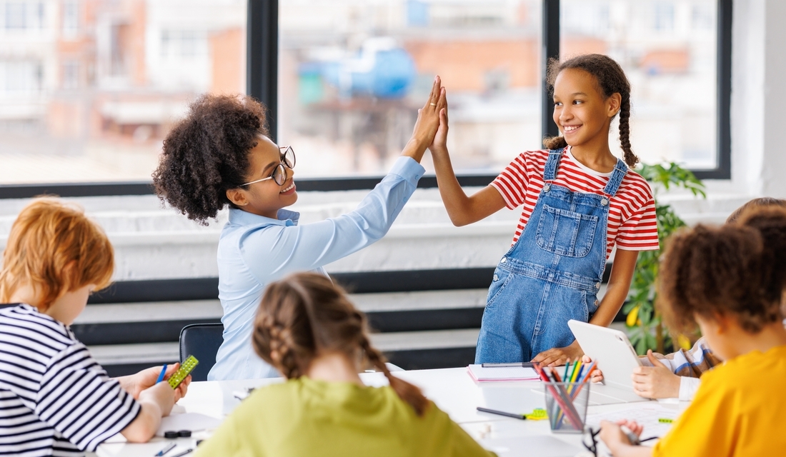 Teacher giving a high-five to a smiling student in a classroom setting, surrounded by other students engaged in learning activities