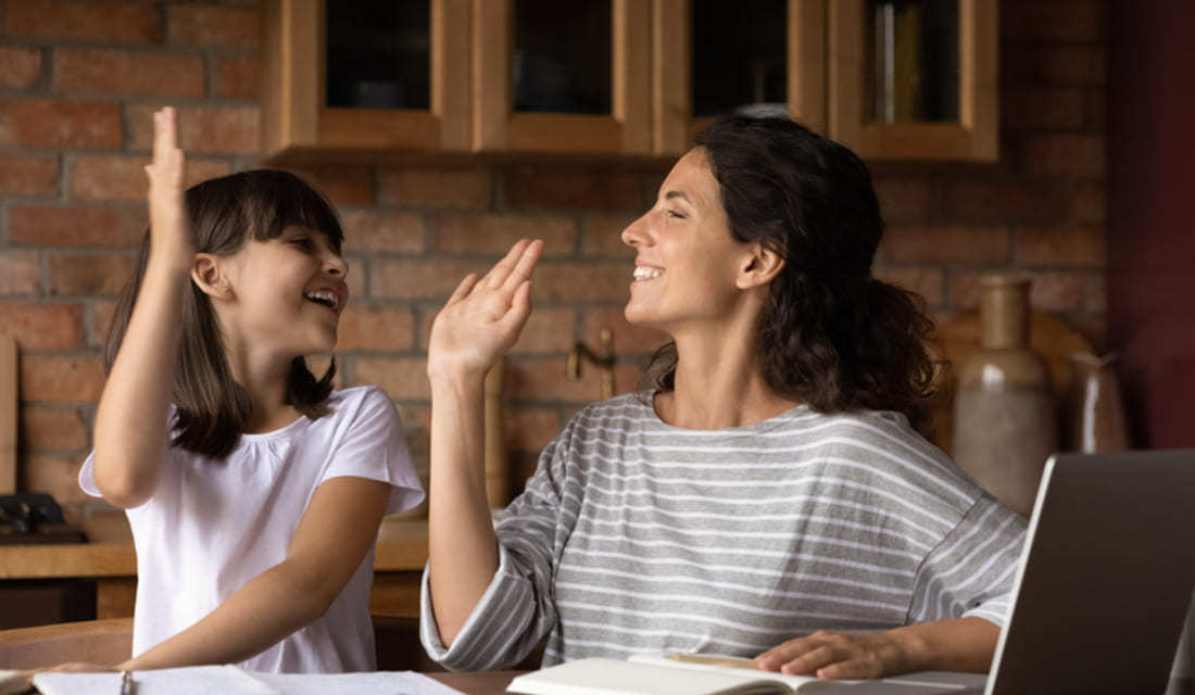 Happy mother and little daughter giving high five, studying math online, doing assignments together, sitting at table with books and laptop at home