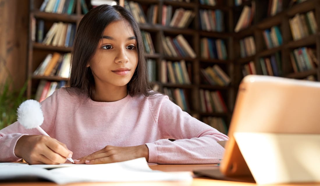 A girl sits at a table with a laptop open and a book beside her, focused on her work.