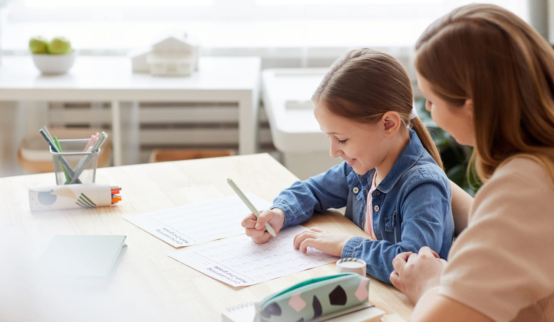 A woman and a young girl sit at a table, focused on writing together with notebooks and pens in front of them.