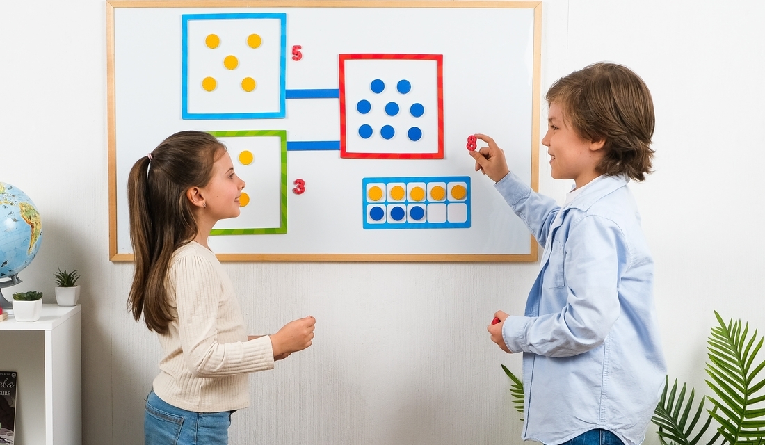 Two children stand in front of a whiteboard filled with various numbers, engaged in a learning activity.