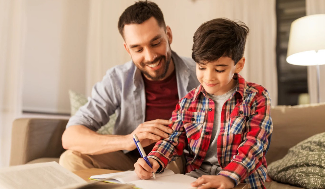 A father and son sit together on a couch, focused on writing in notebooks.