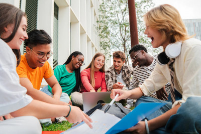 A group of students sitting on grass, collaborating on a laptop in a sunny outdoor setting.