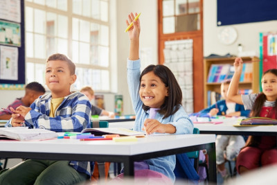 Students in a classroom, sitting at desks with hands raised, indicating their desire to contribute to the discussion.