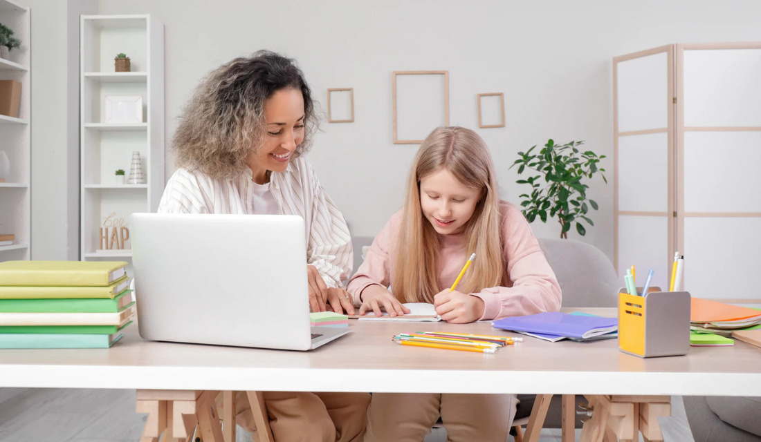 A woman and a girl collaborate at a table, both looking at a laptop placed between them.