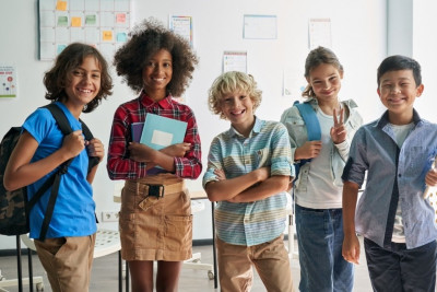 A group of children in a classroom, each wearing backpacks and engaged in conversation.