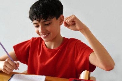 A boy in a red shirt sits at a desk, raising his fist while focused on his homework.