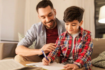 A man and a young boy are seated on a couch, engaged in writing activities with notebooks.