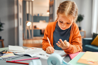 A young girl sits at a table, focused on writing in her notebook with a pencil in hand.