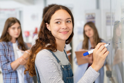 A girl writes on a whiteboard while other students observe and engage in a classroom setting.