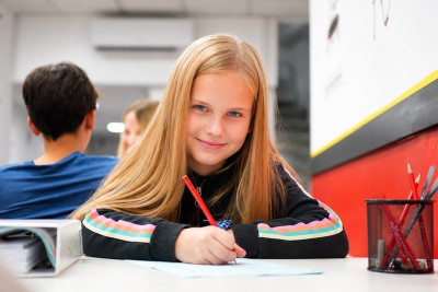 A girl sits at a table, focused on writing in a notebook with a pen in her hand.