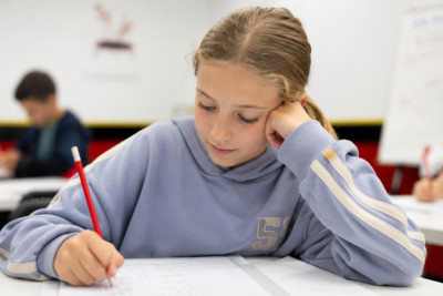 A girl writes in a notebook at her desk, surrounded by classmates engaged in a classroom setting.